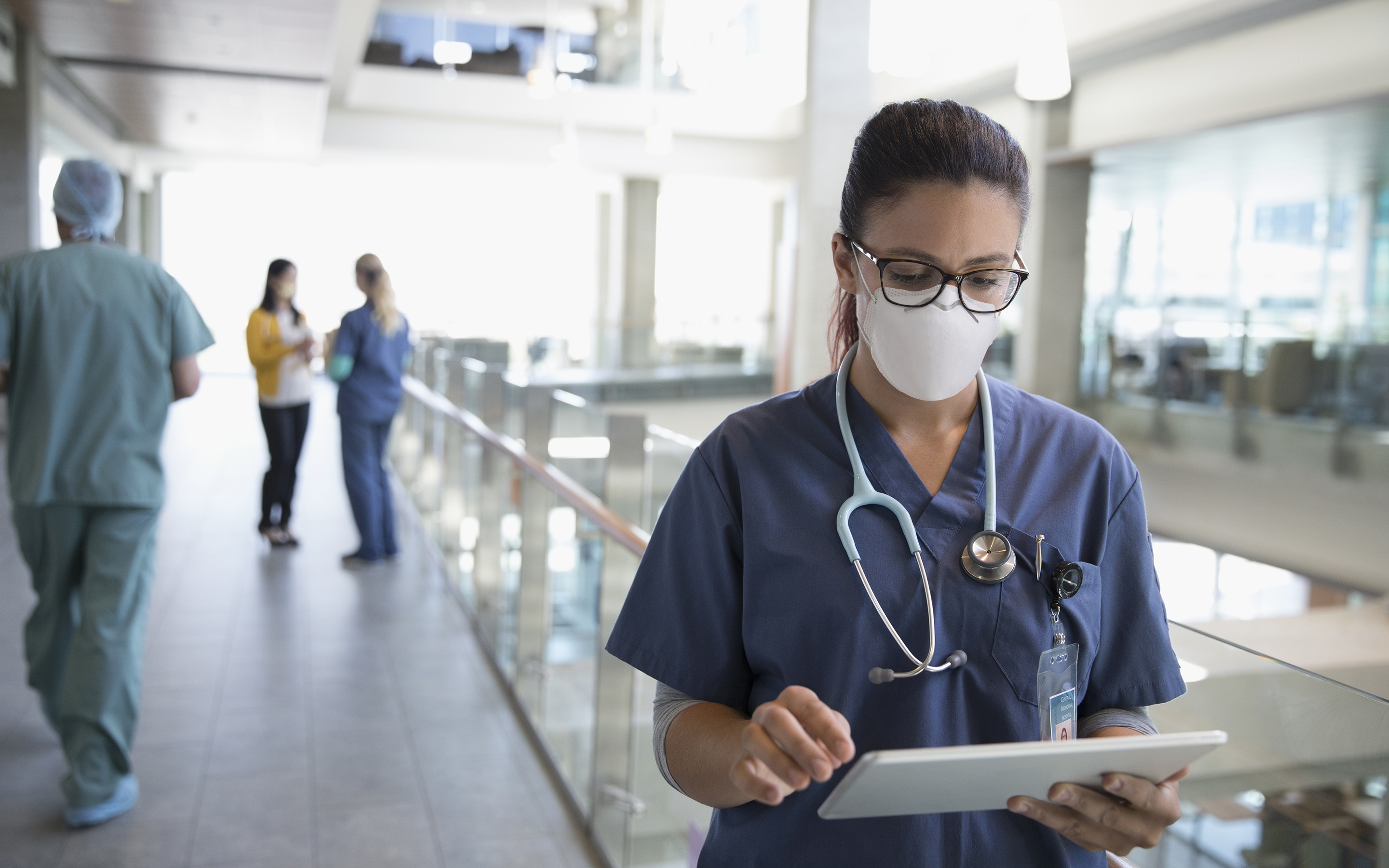 Masked clinician looking at her tablet while standing in a hospital hallway 