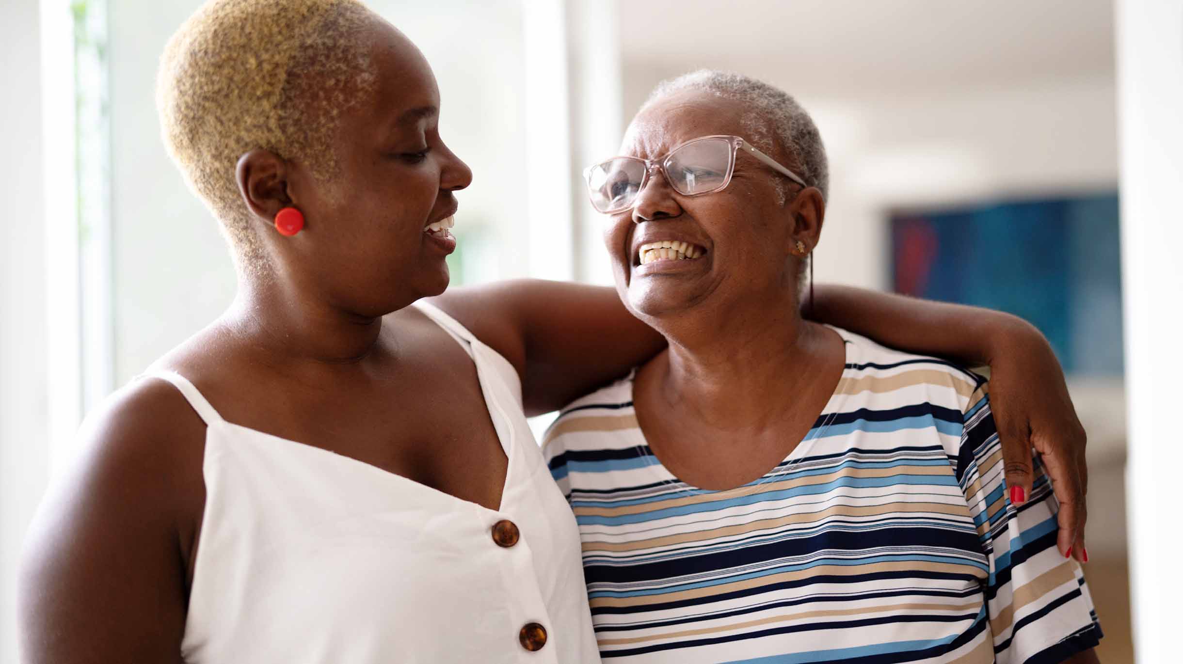 A mother and her adult-aged daughter share an embrace in a bright living room