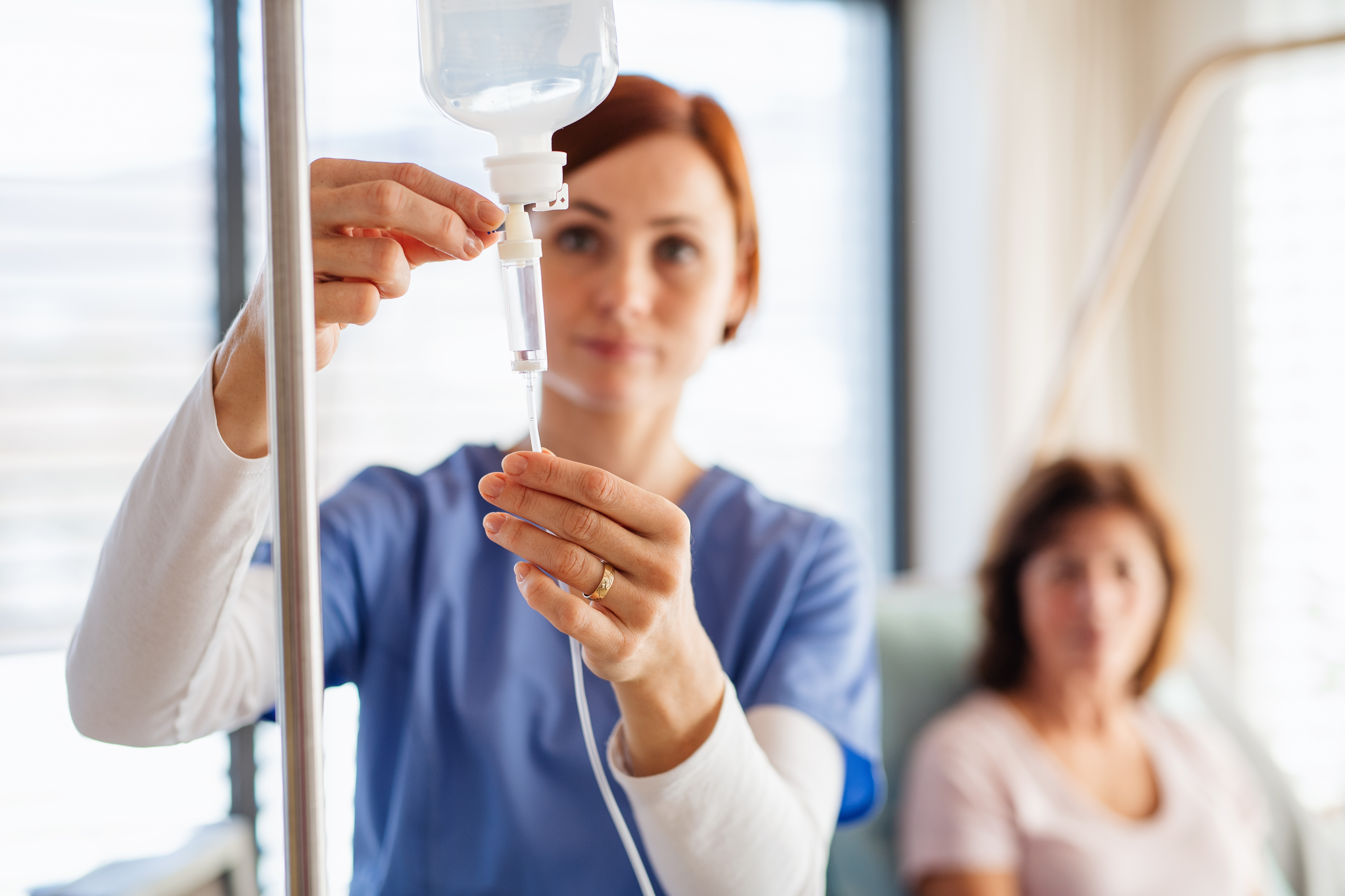 A nurse hanging an infusion bag with a patient sitting in the background